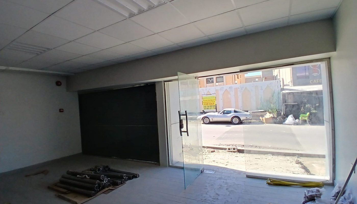 Interior of an empty commercial space with large glass windows overlooking a street, construction materials on the floor, and a closed roller shutter door. This setup is ideal for SEO keywords strategy sessions.