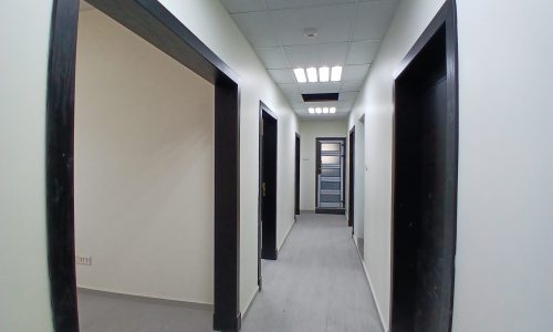 Interior of an office corridor with multiple doorways and a glass door at the end, illuminated by overhead auto draft lights.