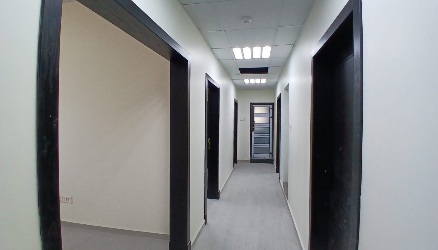 Interior of an office corridor with multiple doorways and a glass door at the end, illuminated by overhead auto draft lights.