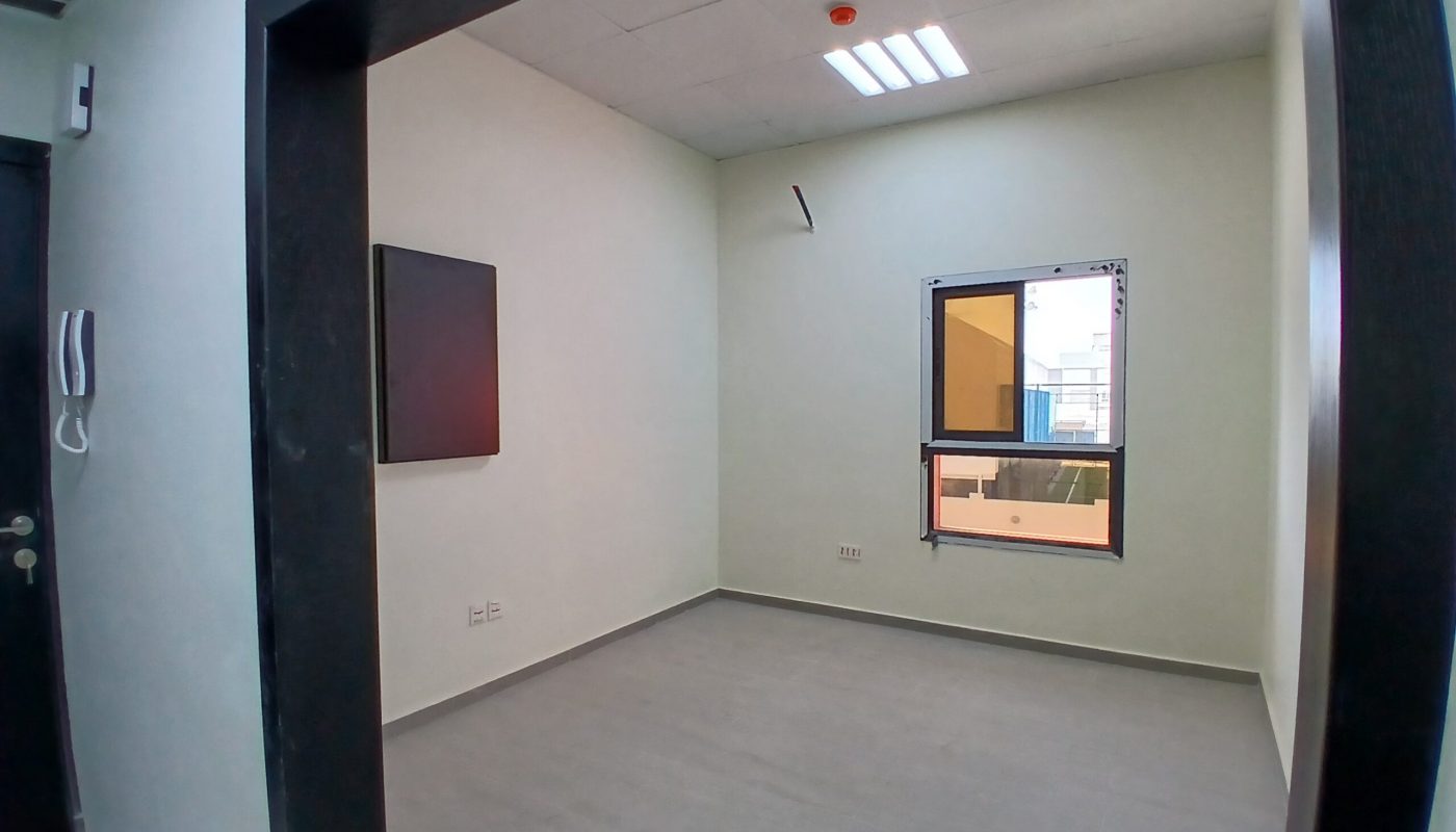 An auto-drafted empty office room with gray carpet, one mounted TV on the left wall, and two windows, one facing out and another smaller one on the adjacent wall.