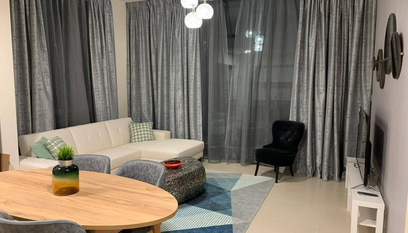 A modern living room with a white sectional sofa, black chair, TV on a white stand, round coffee table, dining table with chairs, and floor-to-ceiling gray curtains.