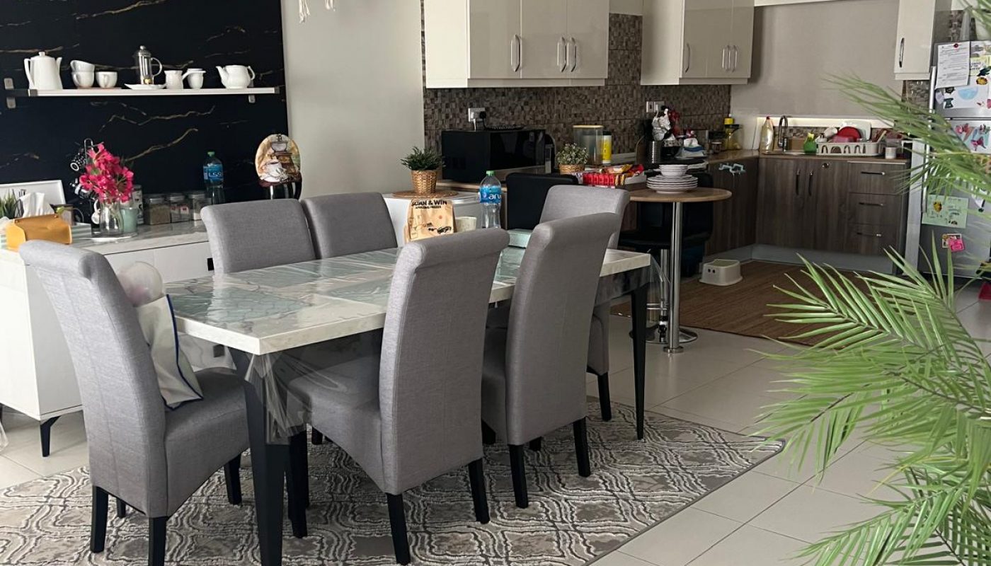 Modern kitchen and dining area with a marble table, six gray chairs, open shelves with dishes, and various items on the countertops and table. A potted plant is in the foreground.