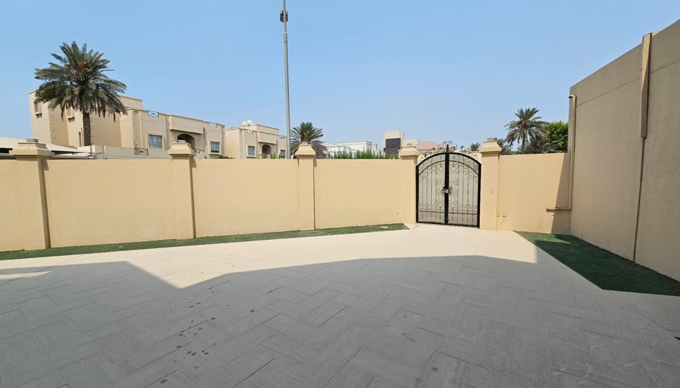 A walled outdoor courtyard with a tiled floor, metal gate, and artificial grass borders, surrounded by beige buildings and palm trees under a clear blue sky.