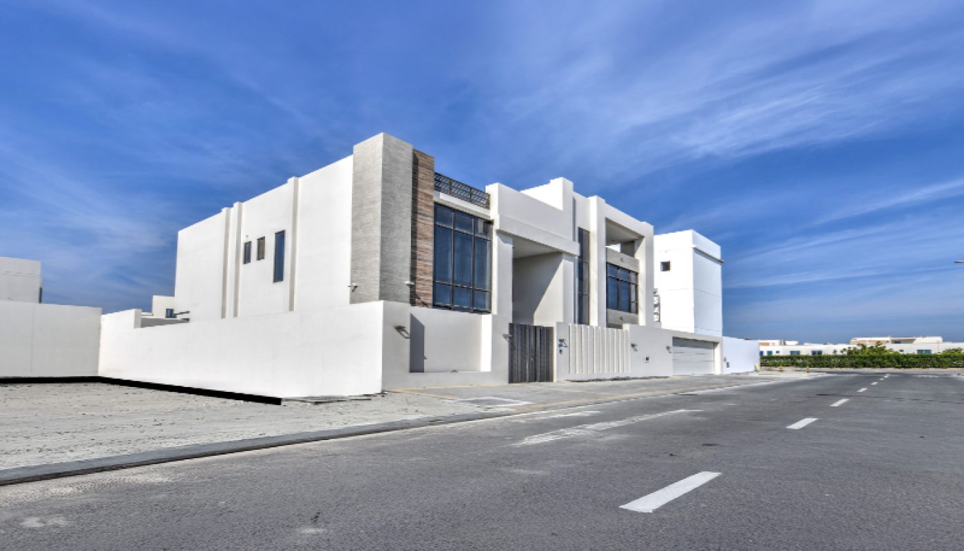 Modern white two-story house with large windows, flat roof, and minimalistic design, situated on a wide, empty street under a clear blue sky.