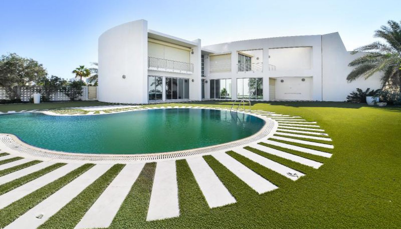 Modern white two-story house with large windows, curved swimming pool, and manicured green lawn, photographed on a sunny day.