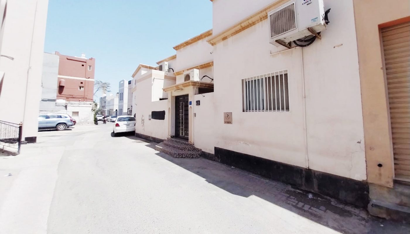 A narrow street with white and beige buildings, barred windows, an air conditioning unit, and parked cars under a clear blue sky.