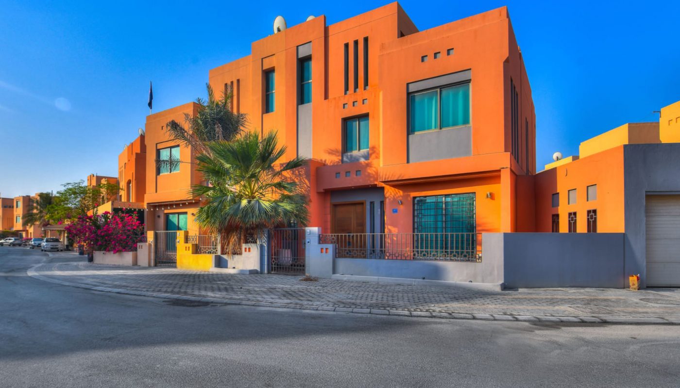 Modern two-story orange building with large windows, palm trees, and a gated entrance on a quiet street under a clear blue sky.