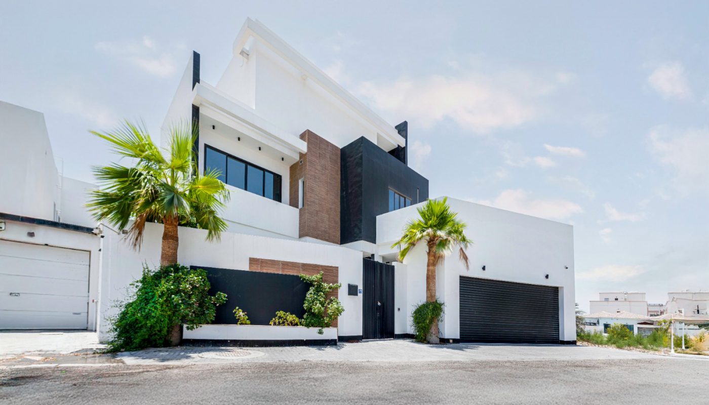 Modern two-story house with white walls, large black garage doors, minimalist design, palm trees, and plants in front, set against a clear sky.