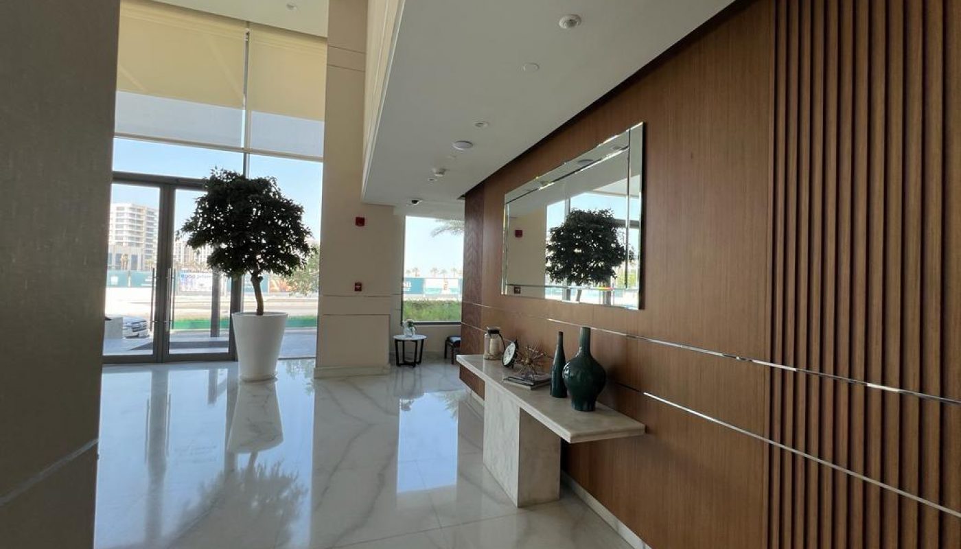 A modern lobby with marble floors, a large mirror on a wooden accent wall, decorative vases on a console table, and a potted tree near glass doors.