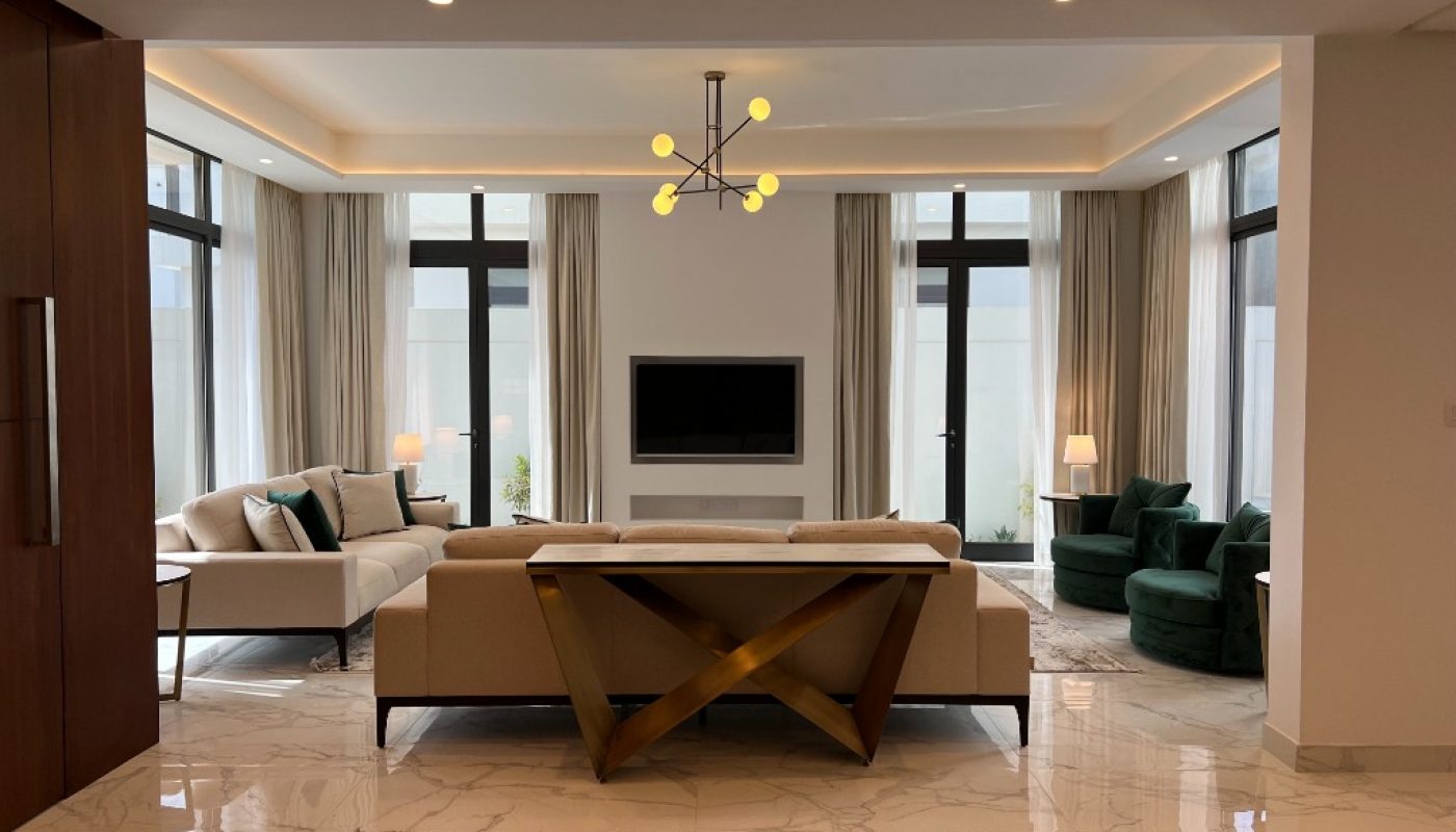 Modern living room with a beige sectional sofa, green chairs, TV on the wall, large windows with curtains, marble floor, and contemporary lighting.