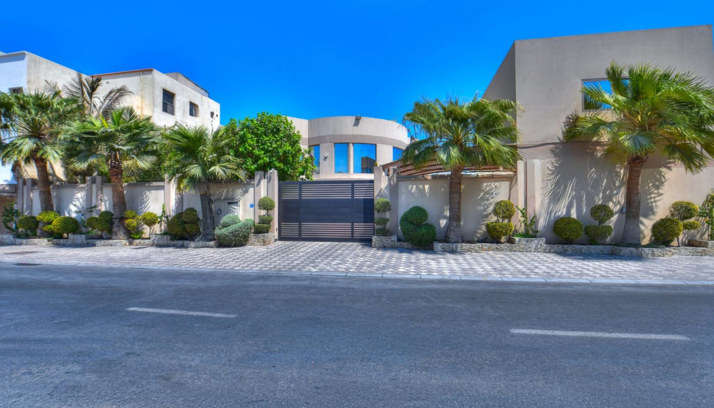 Modern residential building with palm trees and landscaped bushes, viewed from across a paved street under a clear blue sky.