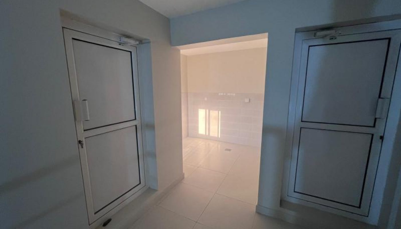 A clean, empty indoor hallway with white tiled floors, off-white walls, and two closed windows on the left and right sides. Natural light shines in from an adjoining room.
