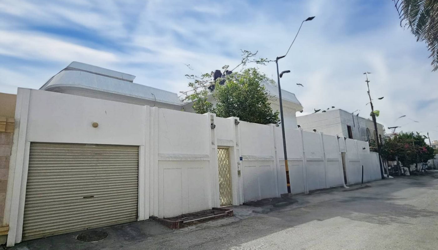 A quiet street lined with white-walled buildings, a closed garage door, and palm trees under a partly cloudy sky.