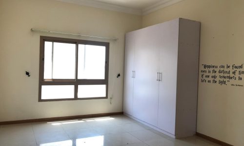 Empty room with a tiled floor, large window, white wardrobe, ceiling light fixture, and a quote on the wall.