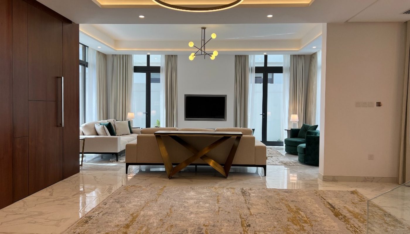 Modern living room with marble floors, neutral-toned furniture, a large area rug, and a central ceiling light fixture. Floor-to-ceiling windows provide natural light from both sides.