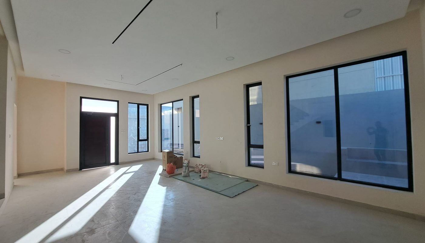 Unfurnished, spacious room with large windows, light-colored walls, and a dark main door. Sunlight streams in, and construction materials are placed on the floor.