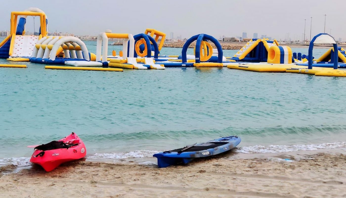 Two kayaks are on the sandy shore in front of an inflatable water obstacle course floating on calm blue water.