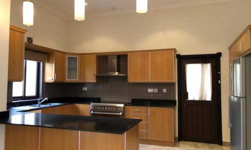 A modern kitchen featuring wooden cabinets, a stainless steel stove and refrigerator, black countertops, tiled backsplash, and hanging pendant lights. A door and window provide natural light.