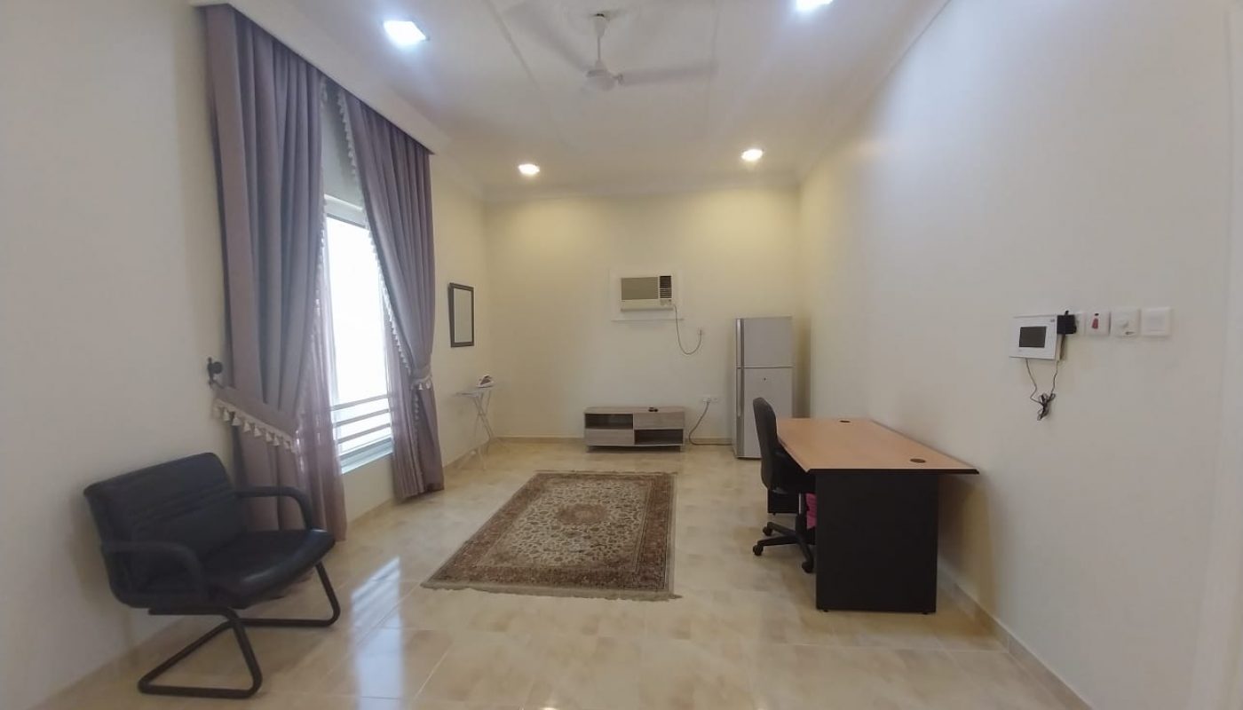 A minimalist room with a desk, chair, black armchair, rug, window with curtains, air conditioner, refrigerator, wall switches, and light-colored tiled floor.