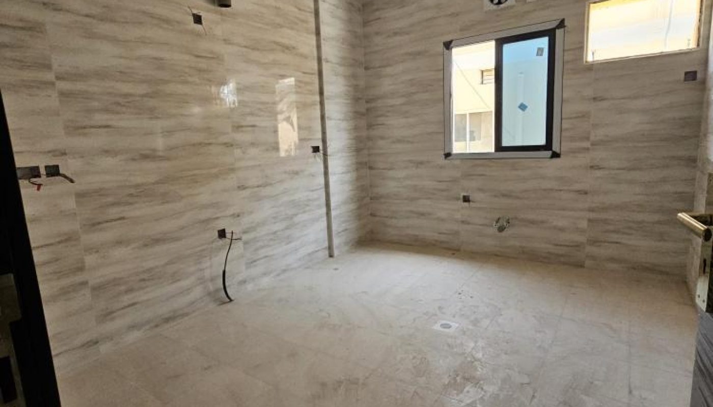 Empty, unfurnished room with tiled walls and floor, a small window, a larger window, and exposed electrical outlets, likely under construction or renovation.