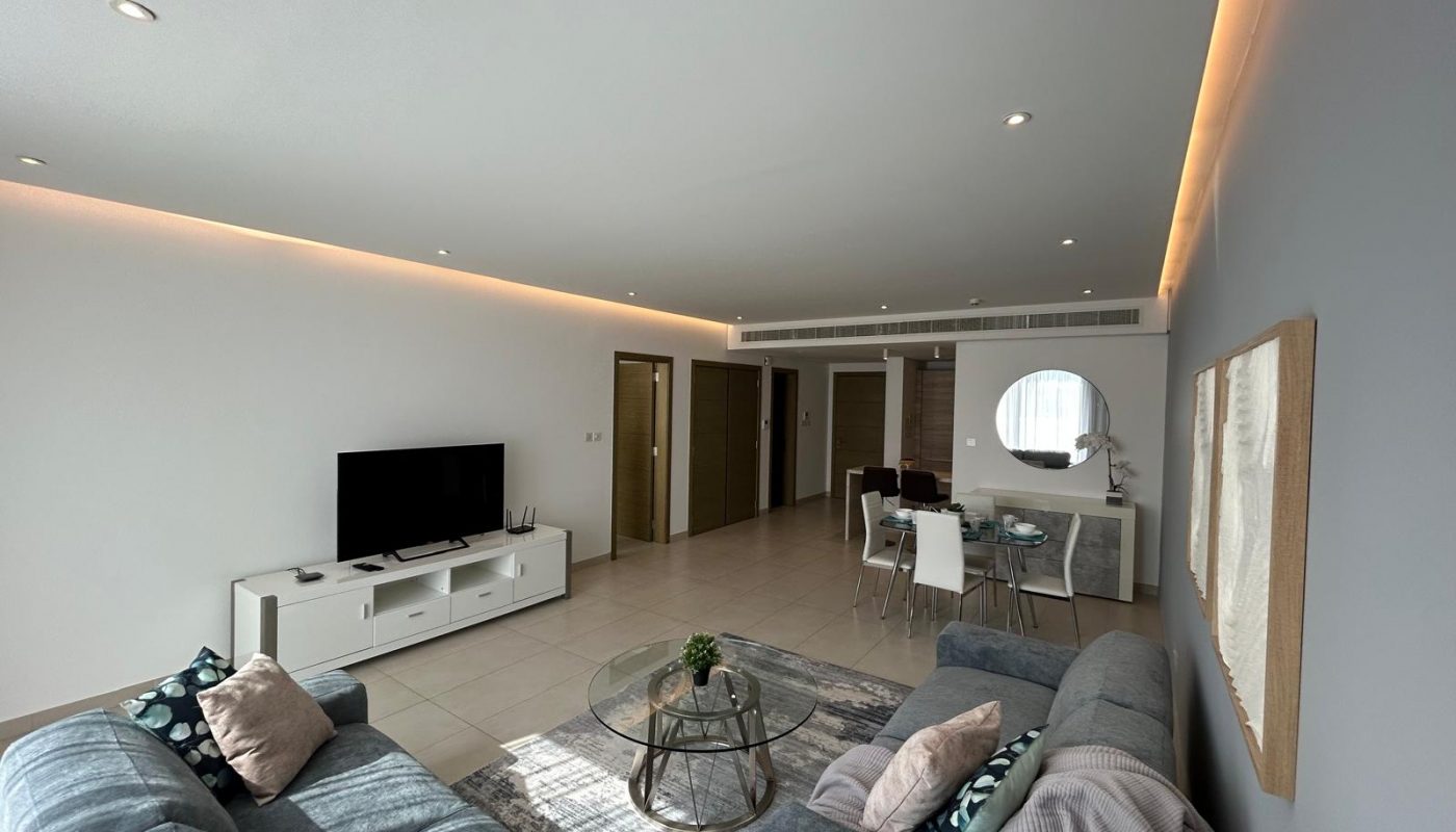 A modern living room with grey sofas, a glass coffee table, a TV on a white stand, and a dining area with a round mirror on the wall. Recessed lighting highlights the ceiling.