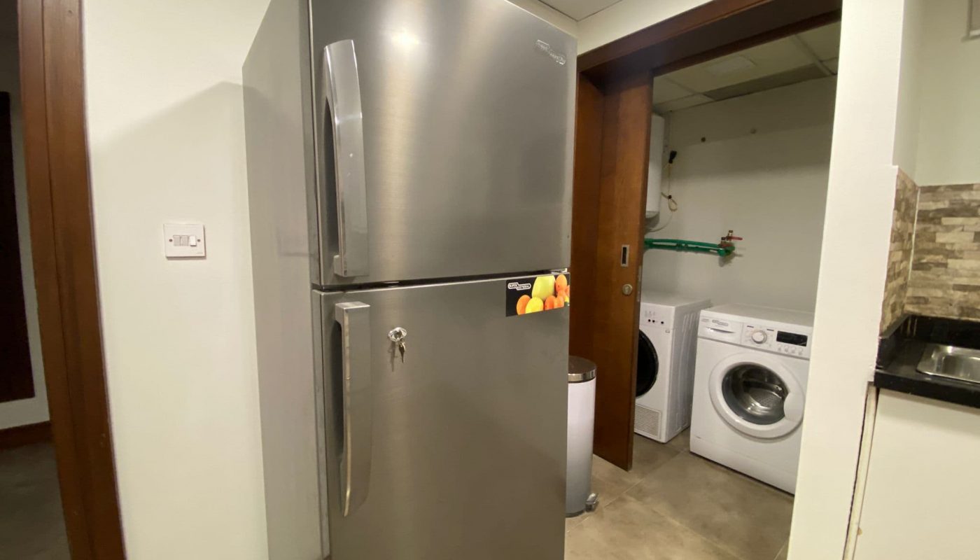 A stainless steel double-door refrigerator is placed next to a laundry area with a washing machine and a countertop in a modern indoor setting.