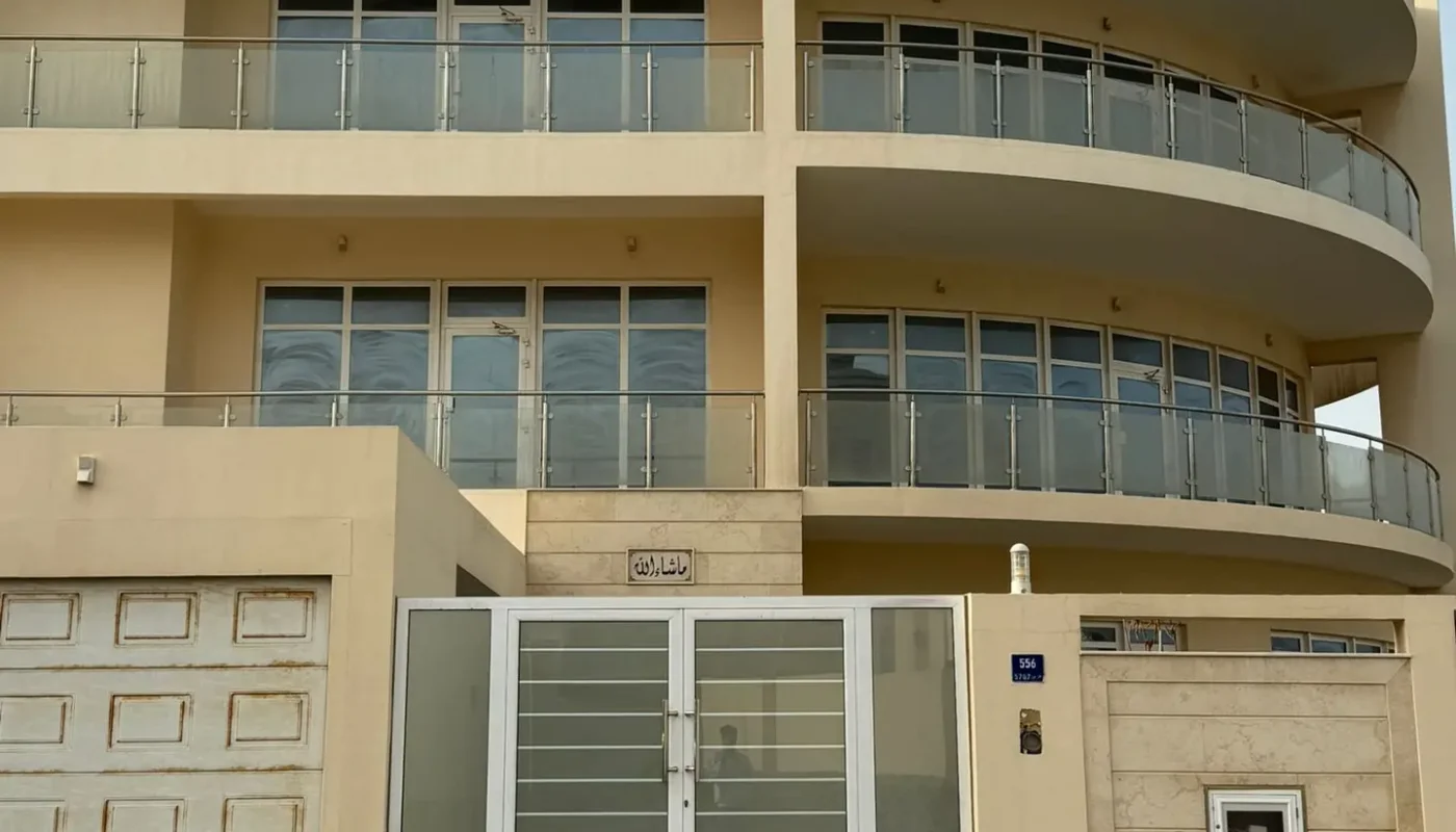 Two-story modern beige building with glass balconies, large windows, a garage, and a double glass gate, viewed from the street.