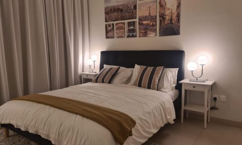 A neatly made bed with white and gray bedding, accented by striped pillows and a brown throw blanket, is showcased in this flat for sale in Diyar Al Muharraq. Two bedside tables with modern lamps are positioned on either side, and framed photos adorn the wall above.