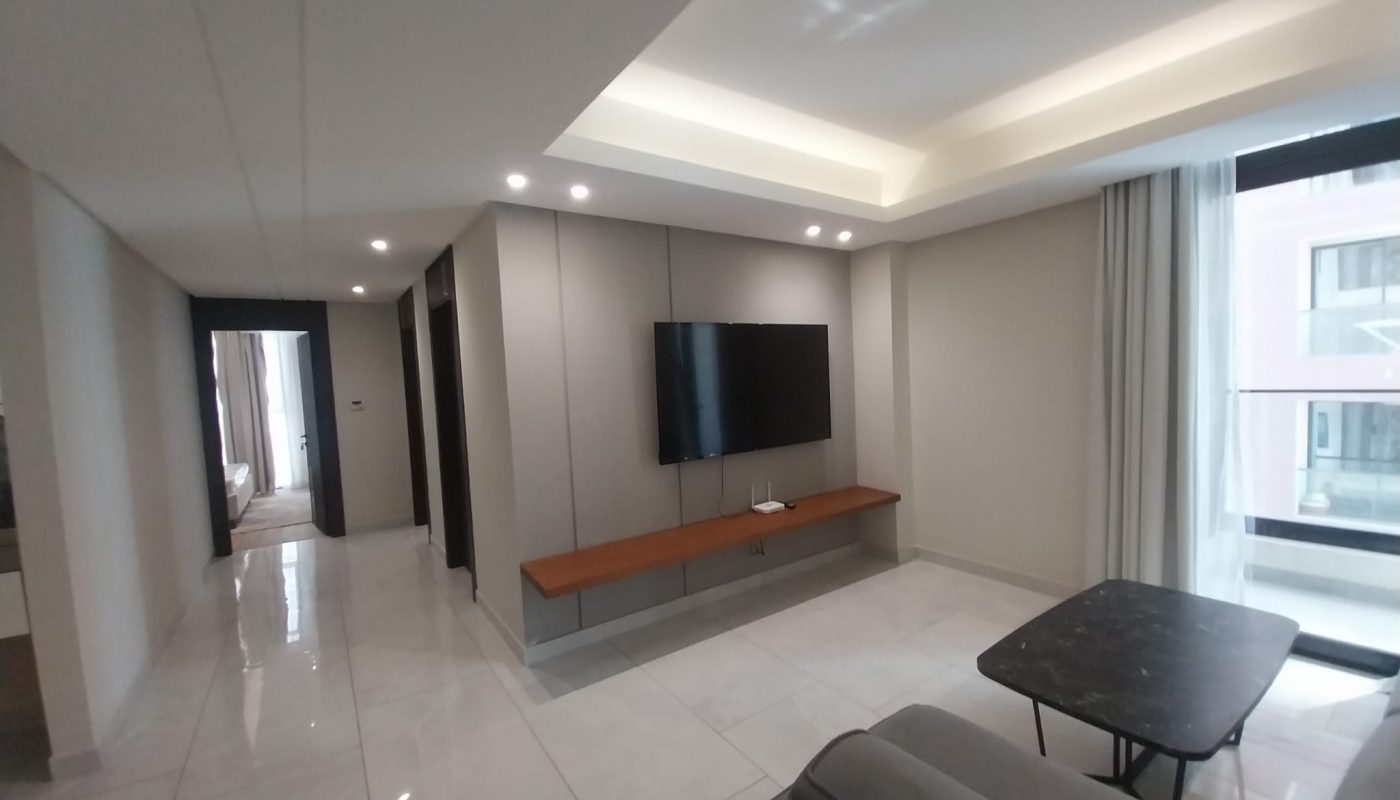 Modern living room with a wall-mounted TV, a wooden shelf, a gray sofa, a black coffee table, and large windows with curtains. Hallway and bedroom visible in the background.