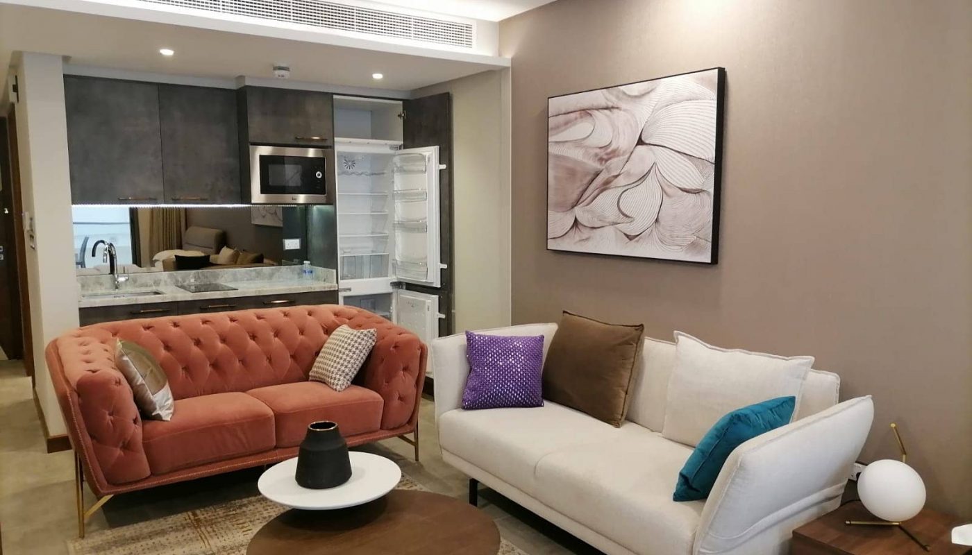 Modern living room with a pink and a white sofa, colorful cushions, round coffee tables, abstract art on the wall, and an open kitchenette with a visible refrigerator.