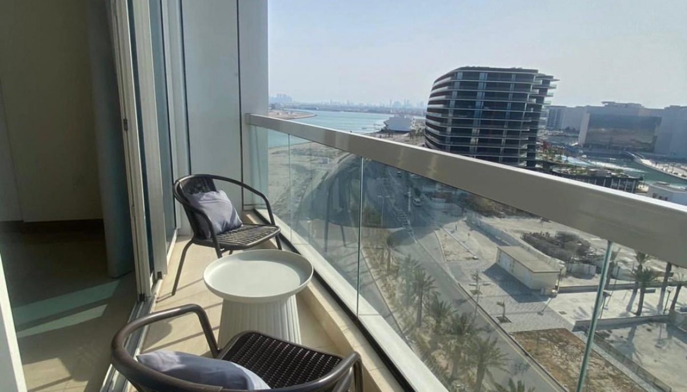 A balcony with two chairs and a small table overlooks a modern cityscape with curved buildings, a road, and distant water under a clear sky.