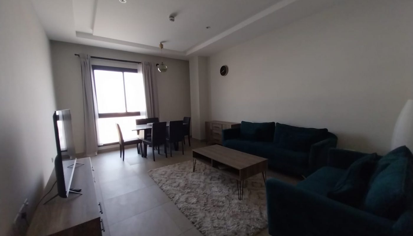 A flat for rent in Hidd features a living room with a dark teal sofa set, a wooden coffee table, a TV on a stand, and a dining table with chairs. Light curtains dress the window, while tiled floors and a cozy rug complete the setup.