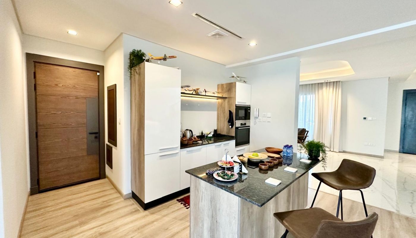 Modern kitchen with wooden flooring, an island with bar stools, built-in appliances, and food items set on the counter; open layout connects to a bright living area.