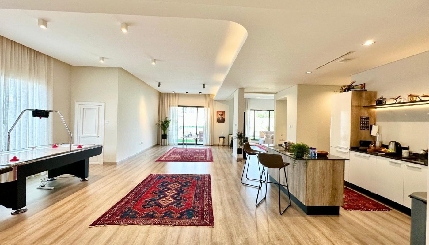 Spacious open-plan living area with kitchen, island seating, air hockey table, two red patterned rugs, large windows, and light wood flooring.