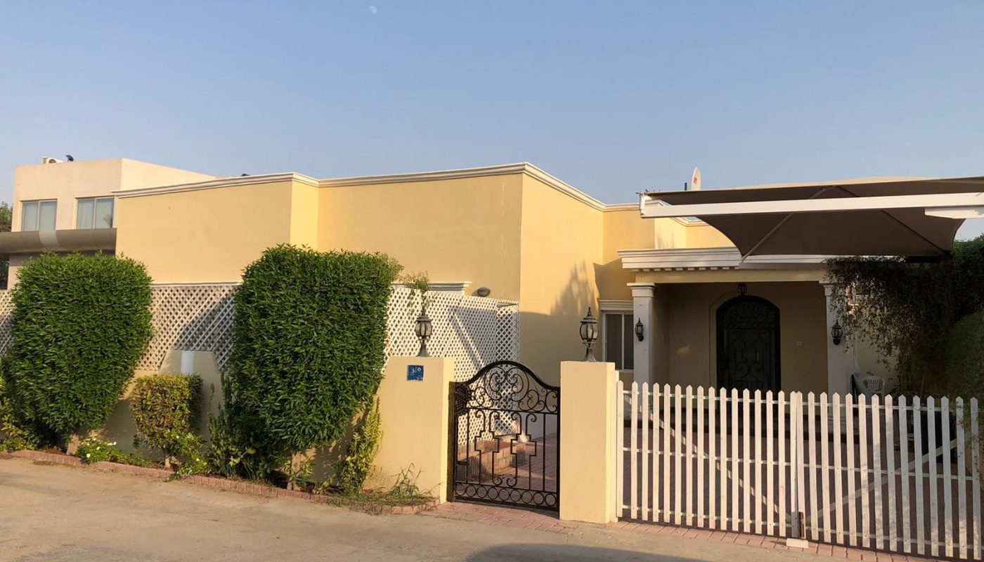 Single-story yellow villa for rent with a white picket fence, a small gate, and green shrubbery under a clear sky at dusk.