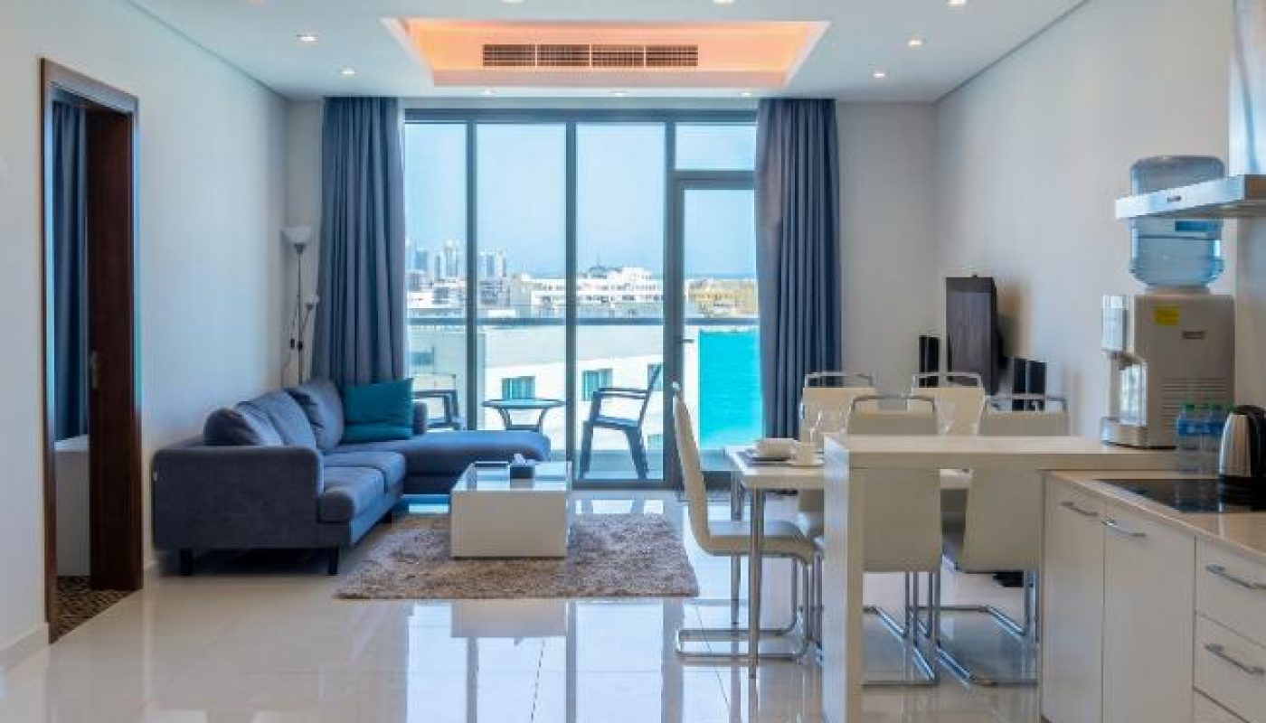 Modern open-plan living room and kitchen with white furniture, tiled floor, large windows, cityscape view, and blue curtains.