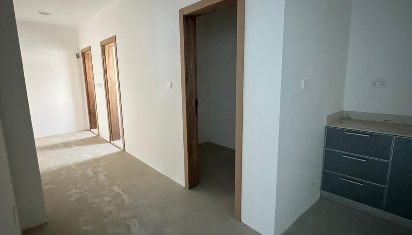 A hallway with unfinished floors, white walls, wooden door frames, a partially open door, and a cabinet with drawers on the right side.