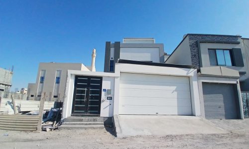 New modern 3BR villa with a white facade and a double garage under a clear blue sky in Malkiya, surrounded by a construction area.