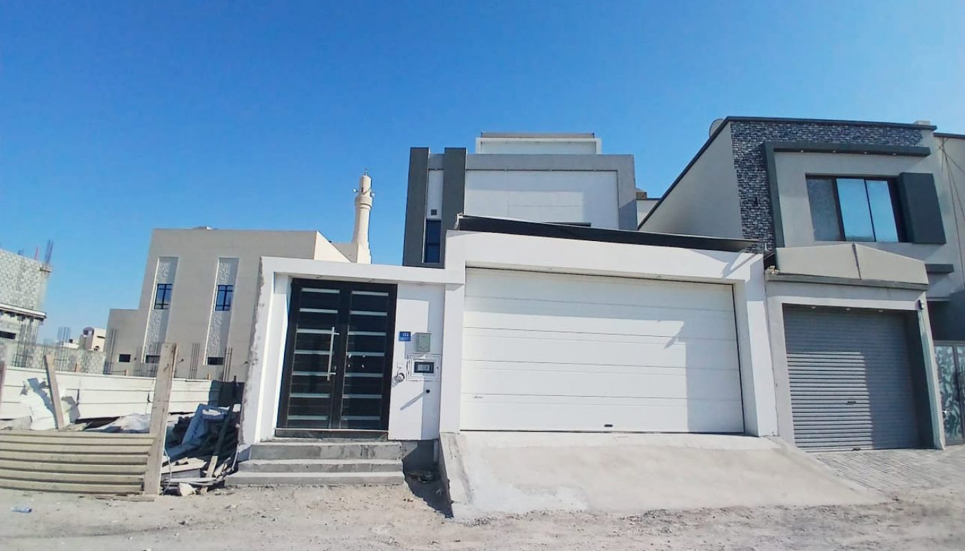New modern 3BR villa with a white facade and a double garage under a clear blue sky in Malkiya, surrounded by a construction area.