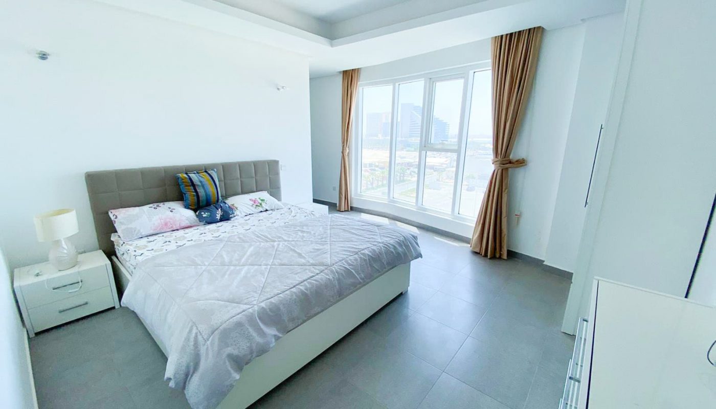 Bright bedroom in a luxury apartment with a large bed, white furniture, and a side table with a lamp. Floor-to-ceiling windows with brown curtains offer a stunning city view. The room features light gray tiles and white walls. Currently on sale in the prestigious Amwaj Towers.