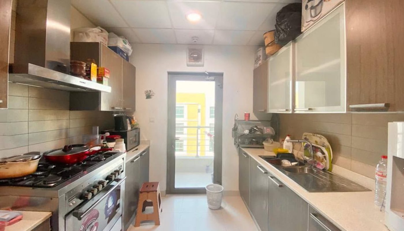 A narrow kitchen with wooden cabinets, a gas stove, and a refrigerator. A doorway at the far end leads to a small balcony with visible railing and natural light streaming in.