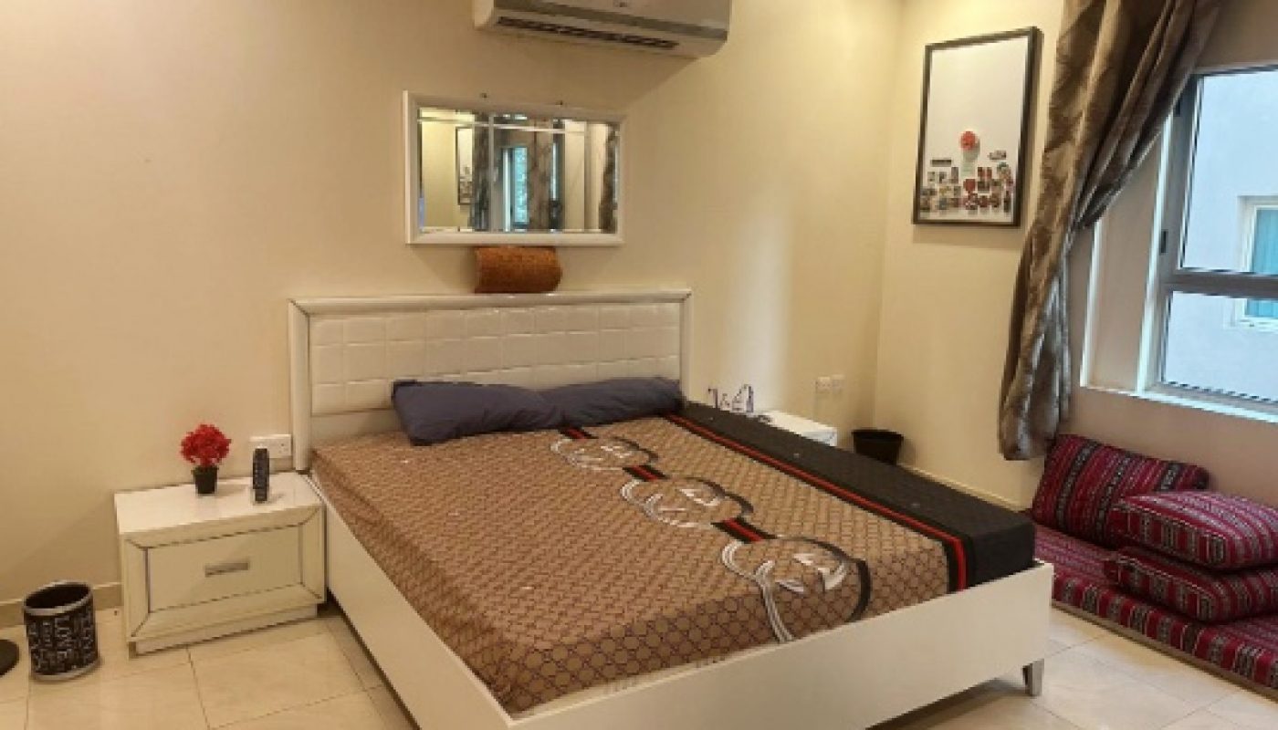 A neatly arranged bedroom with a double bed, bedside table, wall-mounted air conditioner, framed pictures, a window, and brown curtains.