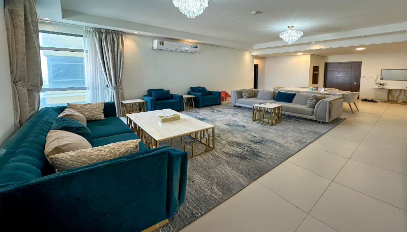 Modern living room with teal and gray sofas, a large rug, two chandeliers, coffee tables, a dining area in the background, and natural light from a window.