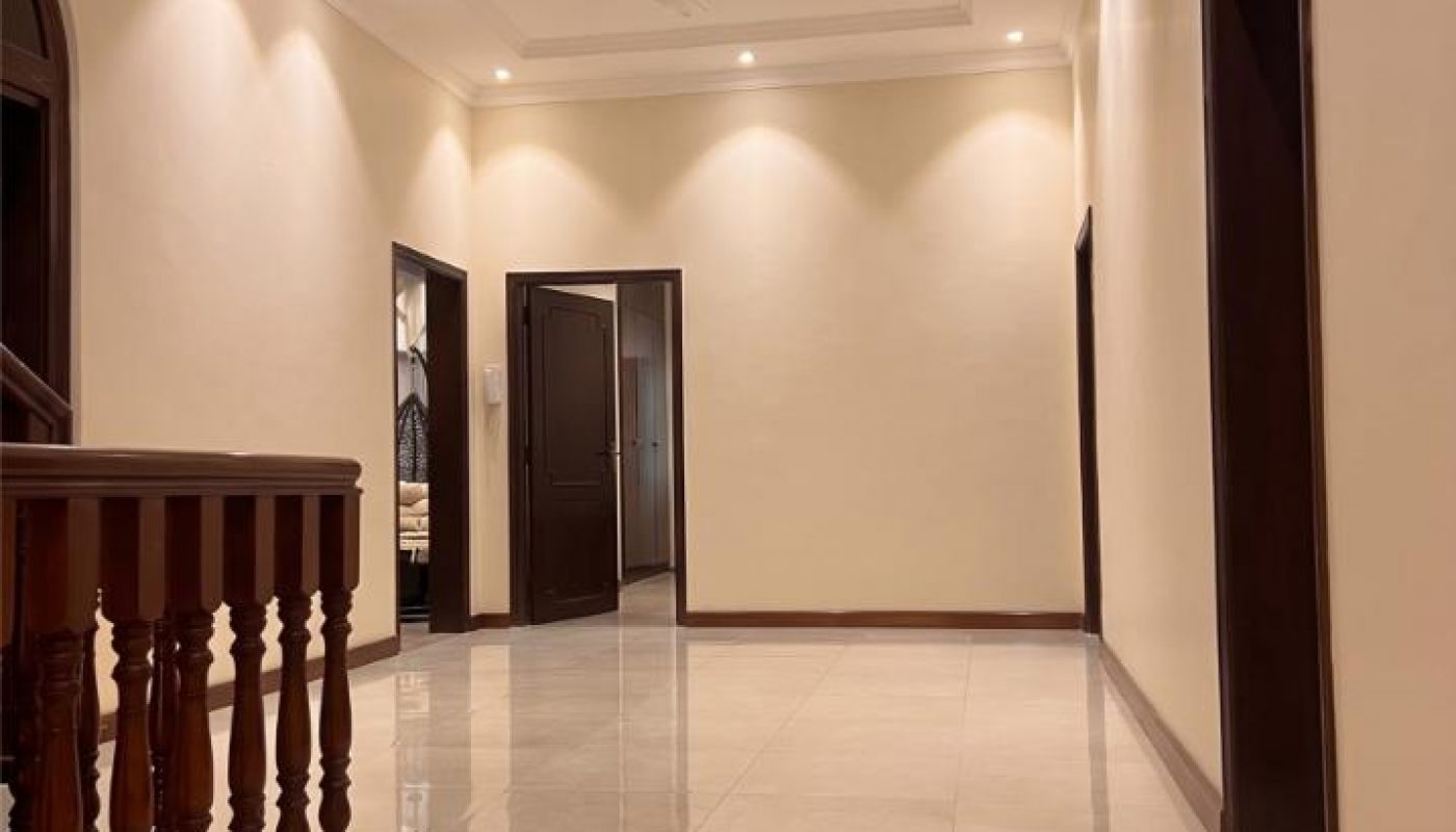 A well-lit hallway with cream-colored walls, tiled floor, ceiling fan, and several open dark-wood doorways.