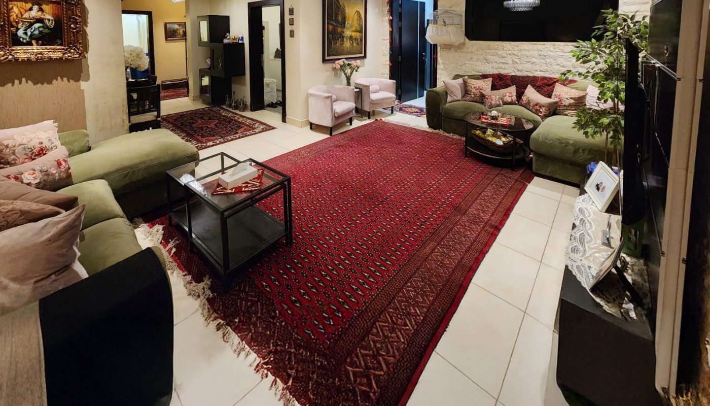 Spacious living room with green sofas, red patterned rug, chandelier lighting, wall art, and a large TV. White tile floor and neutral walls create a bright, elegant atmosphere.