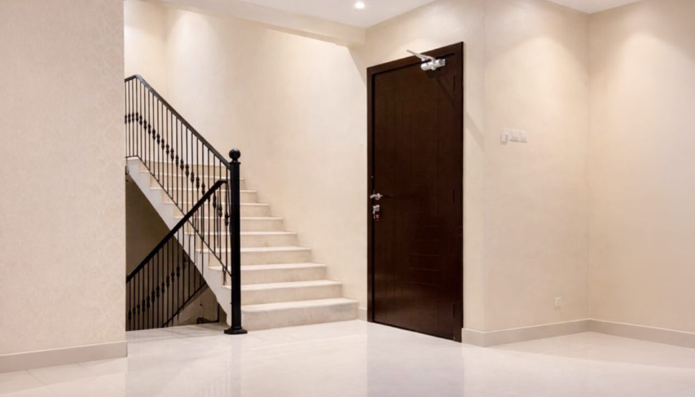 A modern, empty room with white tile flooring, cream walls, a dark wooden door, and a staircase with black railing leading to the upper floor.