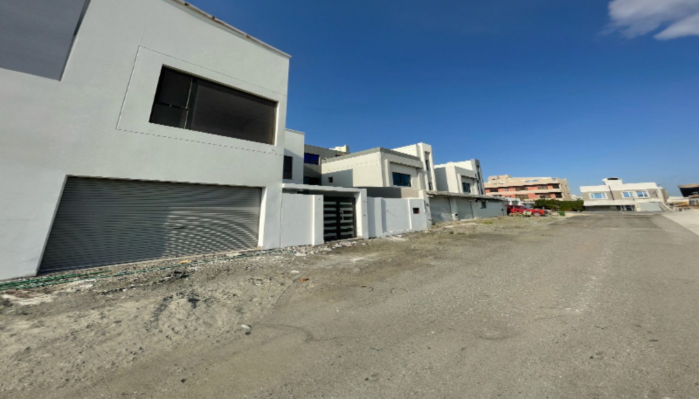 Row of modern, white, two-story buildings with large windows and garages, situated along an unfinished, gravel road under a clear blue sky.