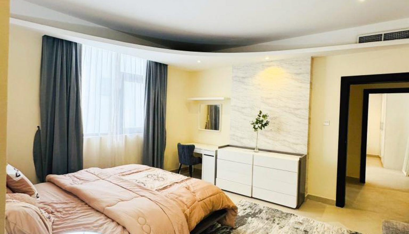 A modern bedroom with a large bed, a dresser with a plant, a desk and chair, gray curtains, and a patterned area rug on a light floor.