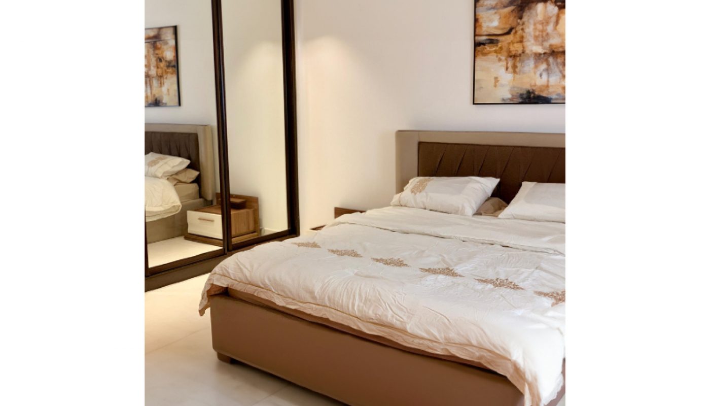 A neatly made bed with a beige headboard, a large mirror closet, white bedding, and abstract artwork on the wall in a minimal, modern bedroom.