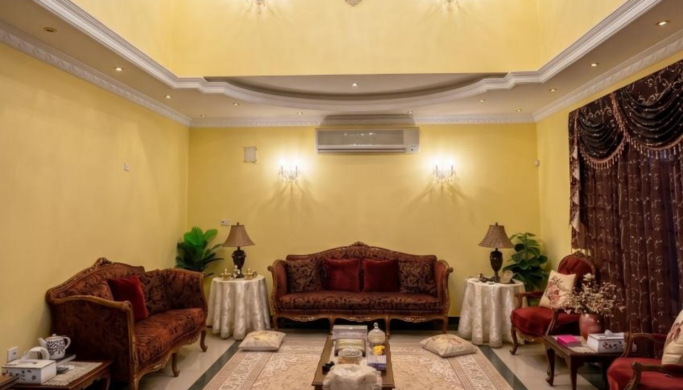 A formal living room with yellow walls, ornate furniture, a large chandelier, a decorative rug, and side tables with lamps and lush curtains.